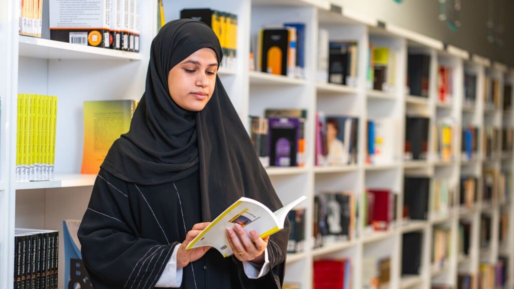Ung kvinne i hijab leser en bok foran bokhyller i et bibliotek